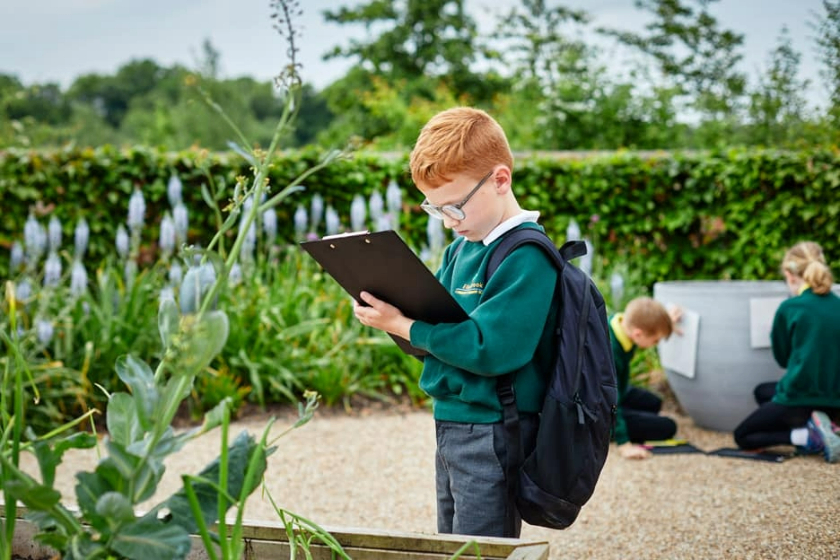 A pupil observes some flowers and writes on a clipboard in a garden