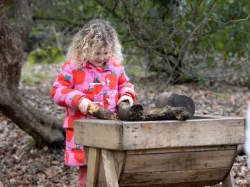 blonde child in brightly coloured coat explores natural materials in a woodland play area